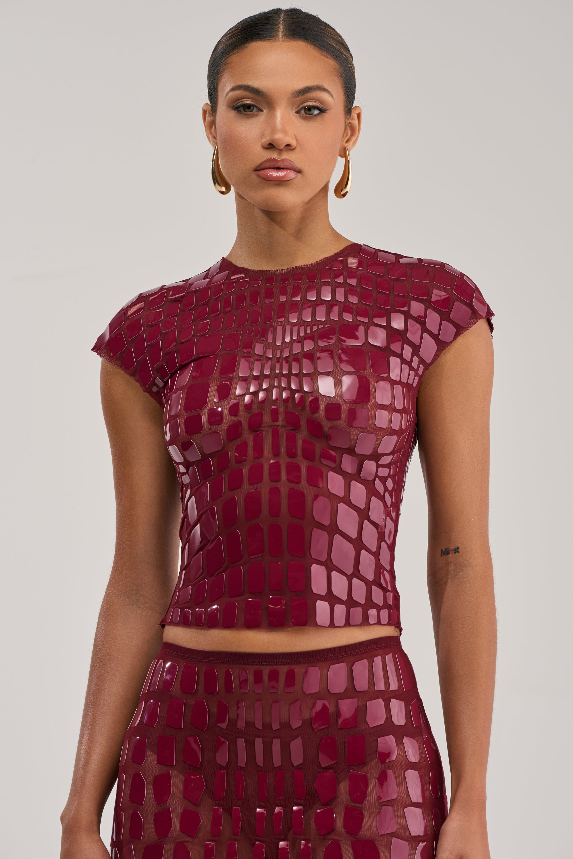A woman in the SEE THROUGH ME TOP IN BURGUNDY, paired with a matching geometric mesh skirt, gold hoop earrings, and slicked-back hair stands against a plain light background.