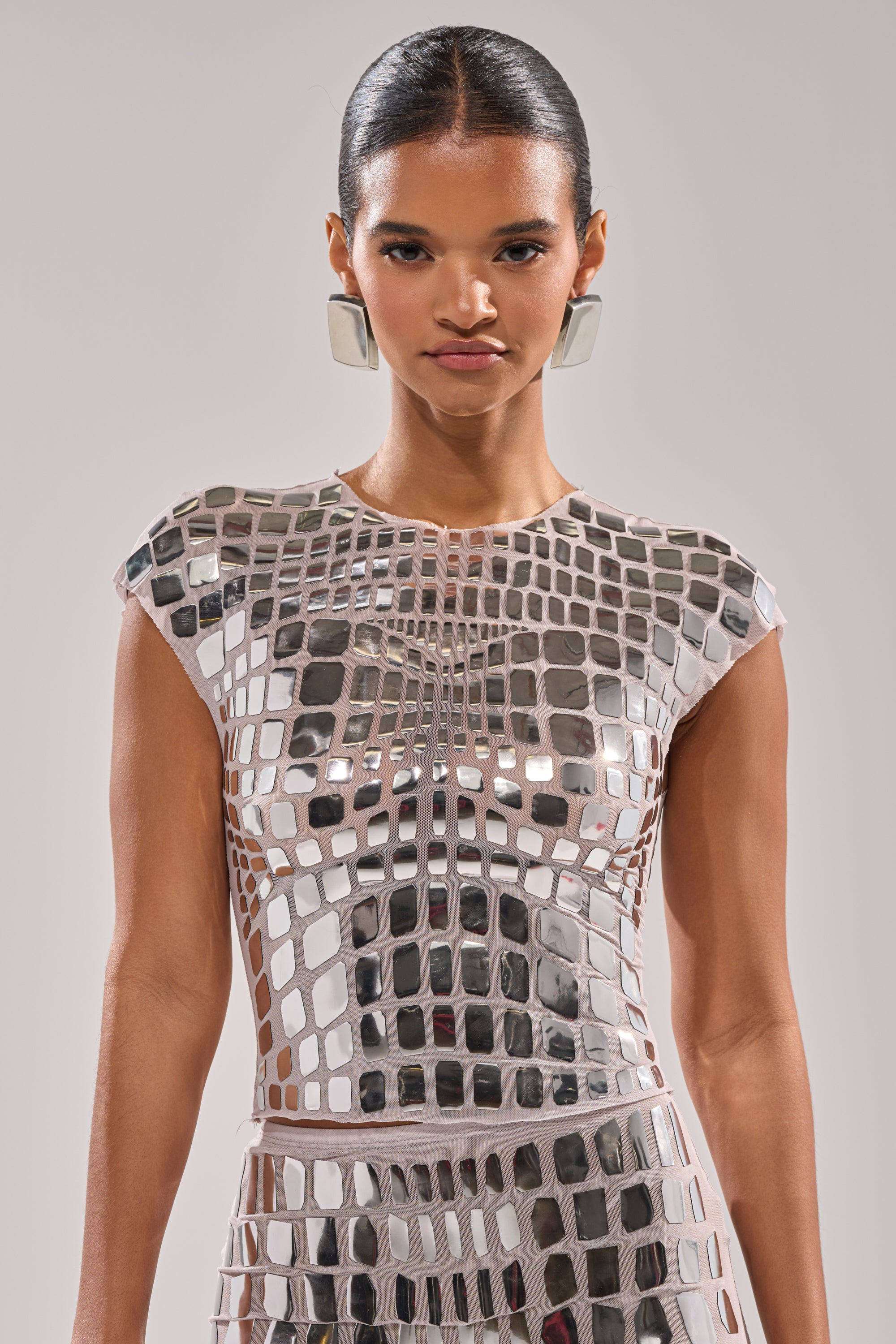A woman with slicked-back hair models the CHAMPAGNE POPPIN BLOUSE, a fitted top adorned with reflective square mirror-like tiles, and pairs it with large geometric earrings against a plain light background.