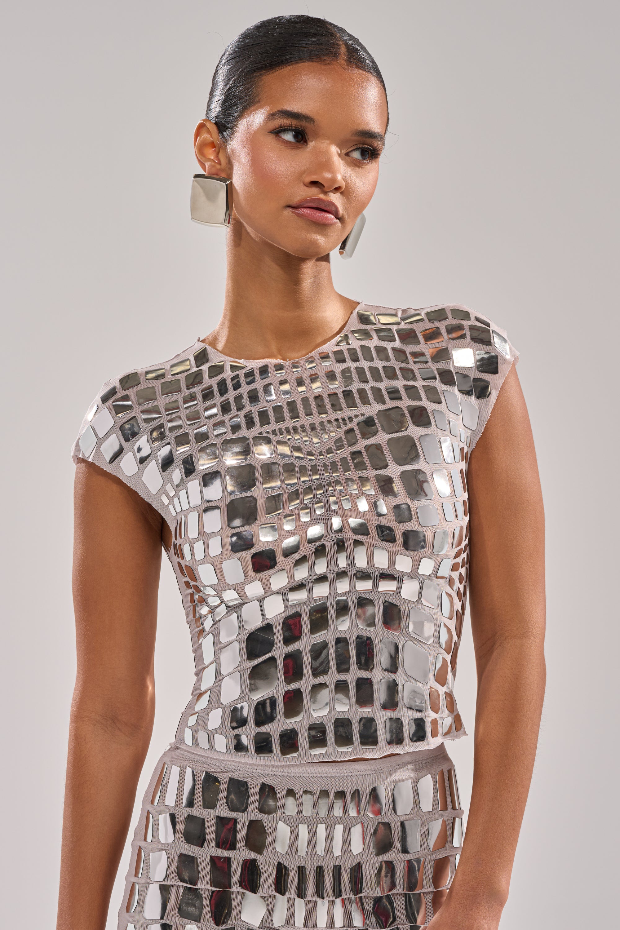 A woman with slicked-back hair models the CHAMPAGNE POPPIN BLOUSE, a mirrored top covered in reflective square tiles, paired with geometric silver earrings against a neutral background.
