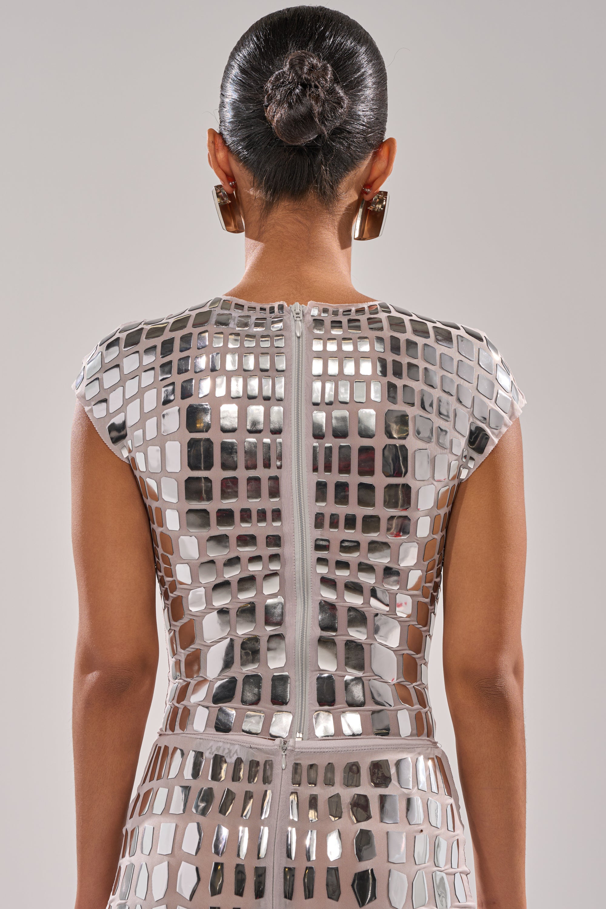 A person with dark hair in a neat bun wears the CHAMPAGNE POPPIN BLOUSE, shimmering with reflective silver squares, paired with large square earrings. The futuristic look is captured from behind against a plain background.