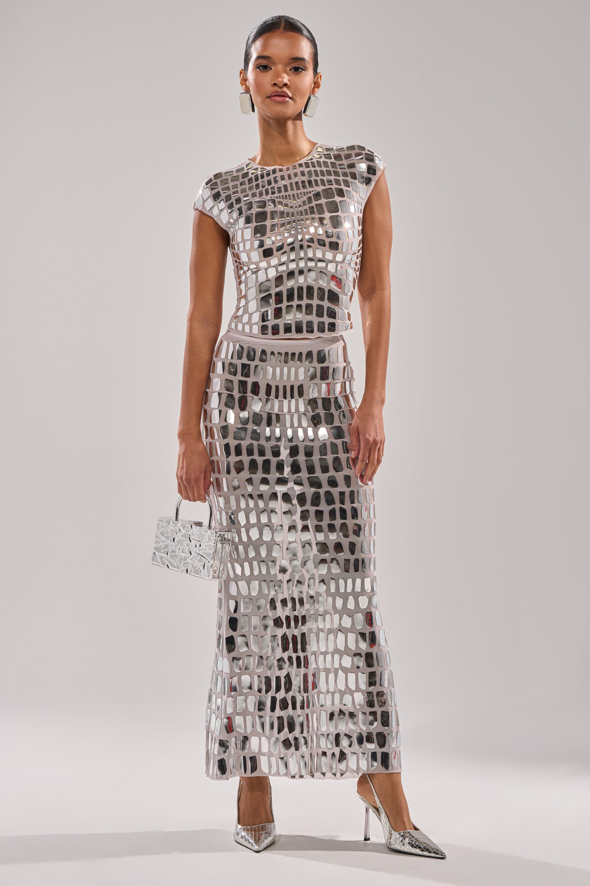 A woman stands confidently in the CHAMPAGNE POPPIN BLOUSE with geometric cutouts, holding a small silver handbag and wearing pointed silver high heels against a plain light background.