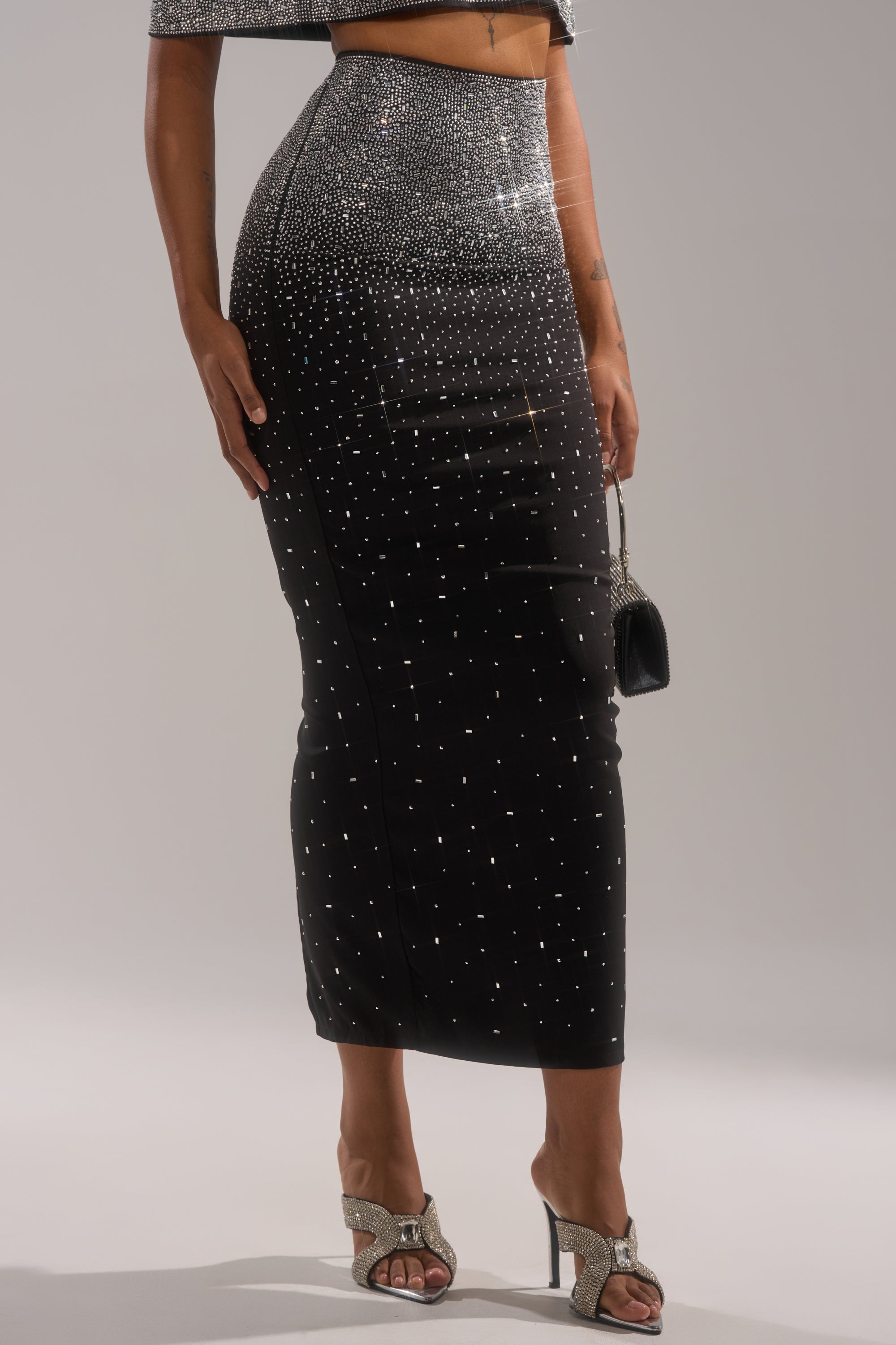 A person wearing the LOVE THIS LIFE MAXI SKIRT—a sparkling black rhinestone skirt with silver embellishments—paired with a matching crop top, silver bow heels, and a small black purse.