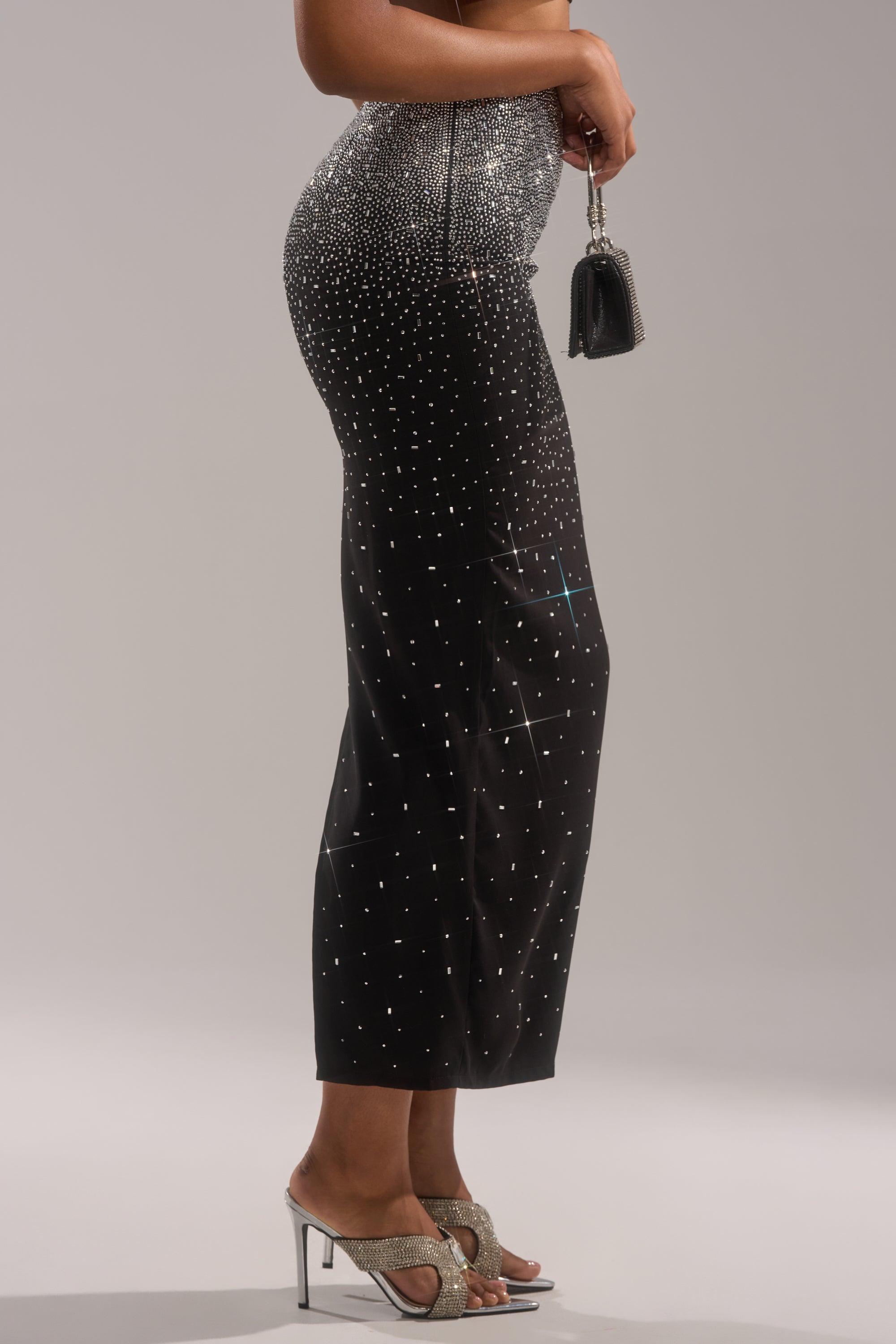 A person stands in profile wearing the LOVE THIS LIFE MAXI SKIRT, featuring a black fabric with a white dotted pattern, paired with silver high-heeled sandals and a small black handbag against a plain light background—effortlessly chic.