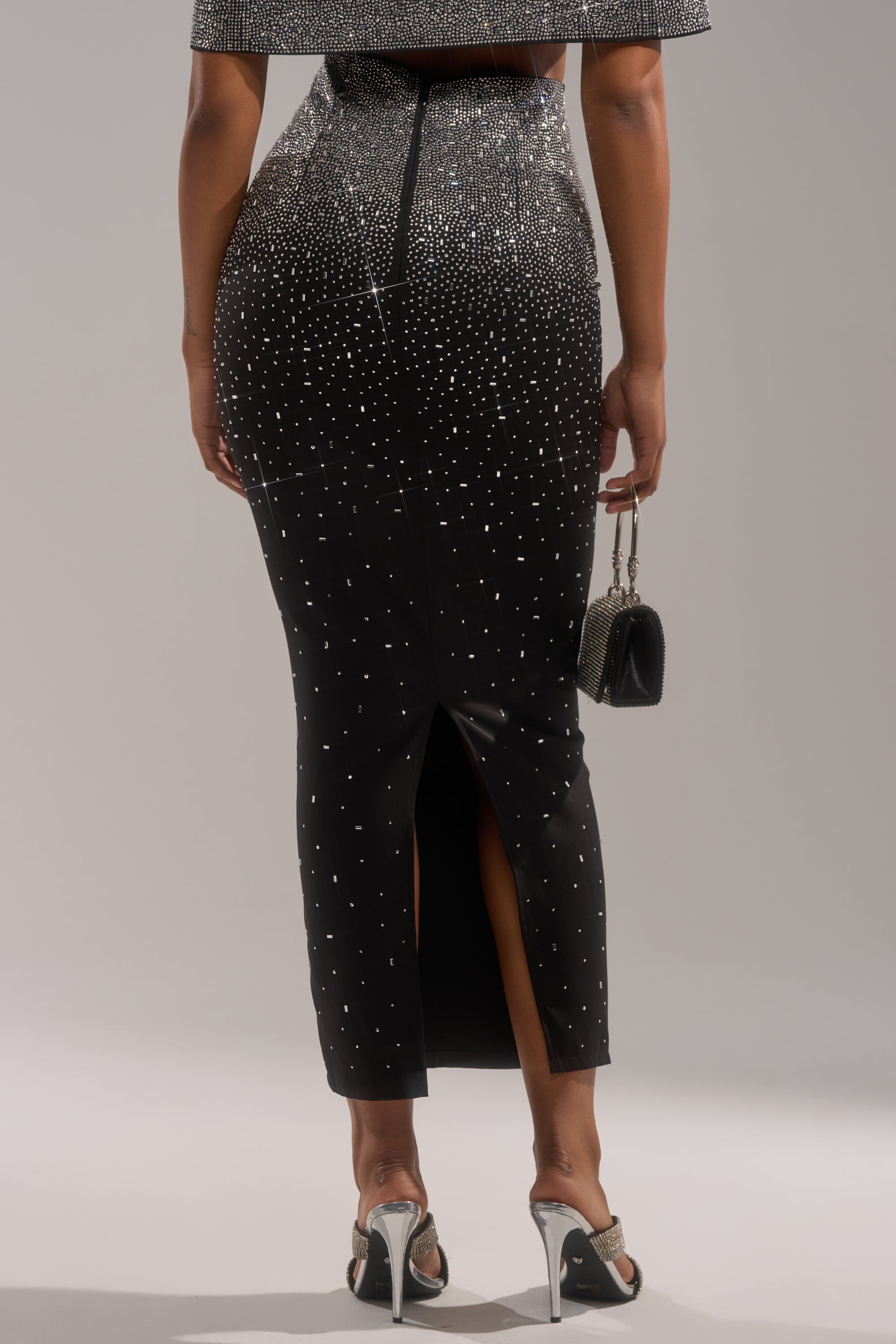 A person is shown from the back wearing the LOVE THIS LIFE MAXI SKIRT, an ankle-length piece with sparkling rhinestone embellishments and a stylish back slit, paired with silver high heels and a glittery handbag.