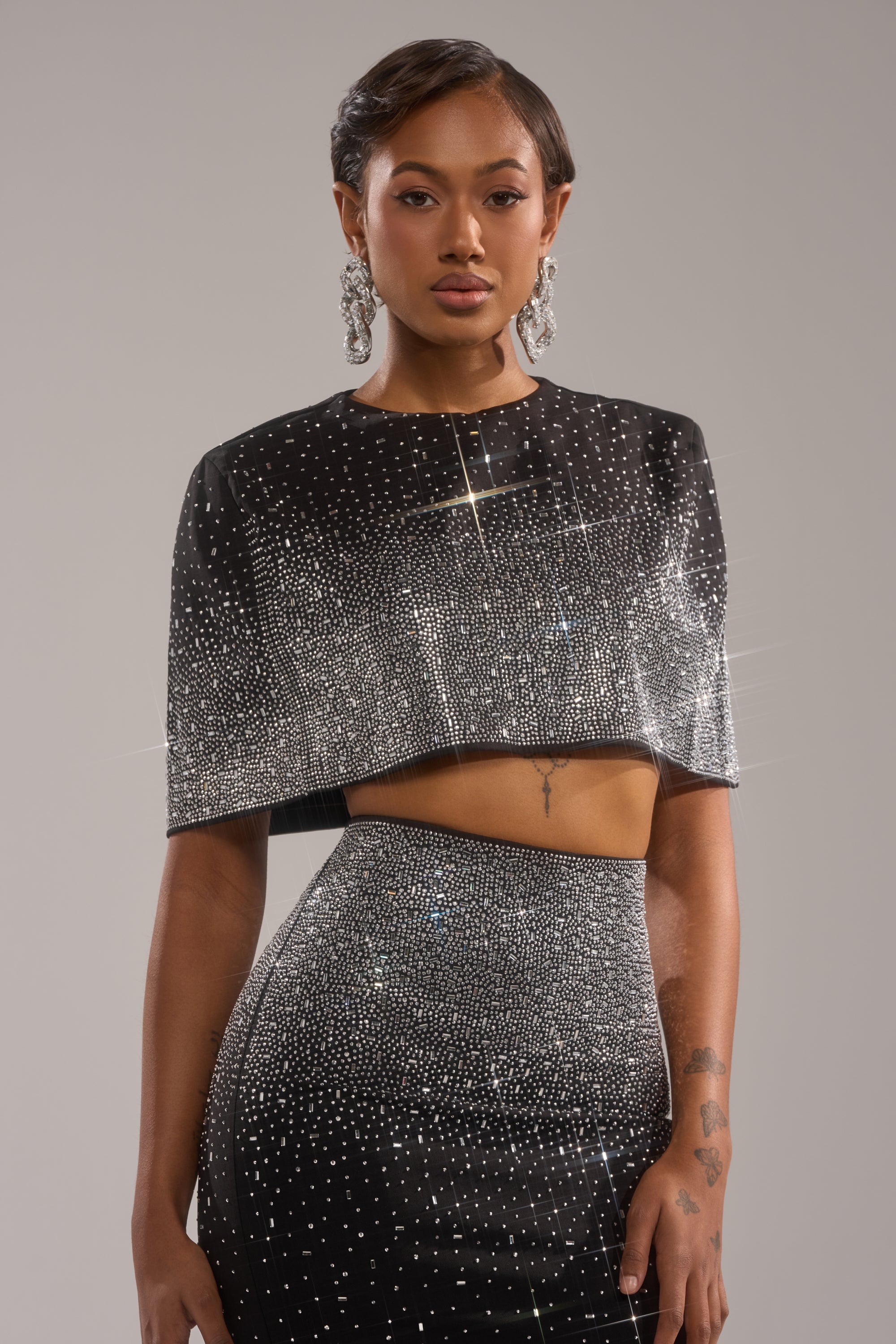 A woman wearing the LOVE THIS LIFE BLOUSE and a matching skirt poses against a plain backdrop. She sports short, styled hair, bold earrings, and has visible tattoos on her left hand and right forearm.