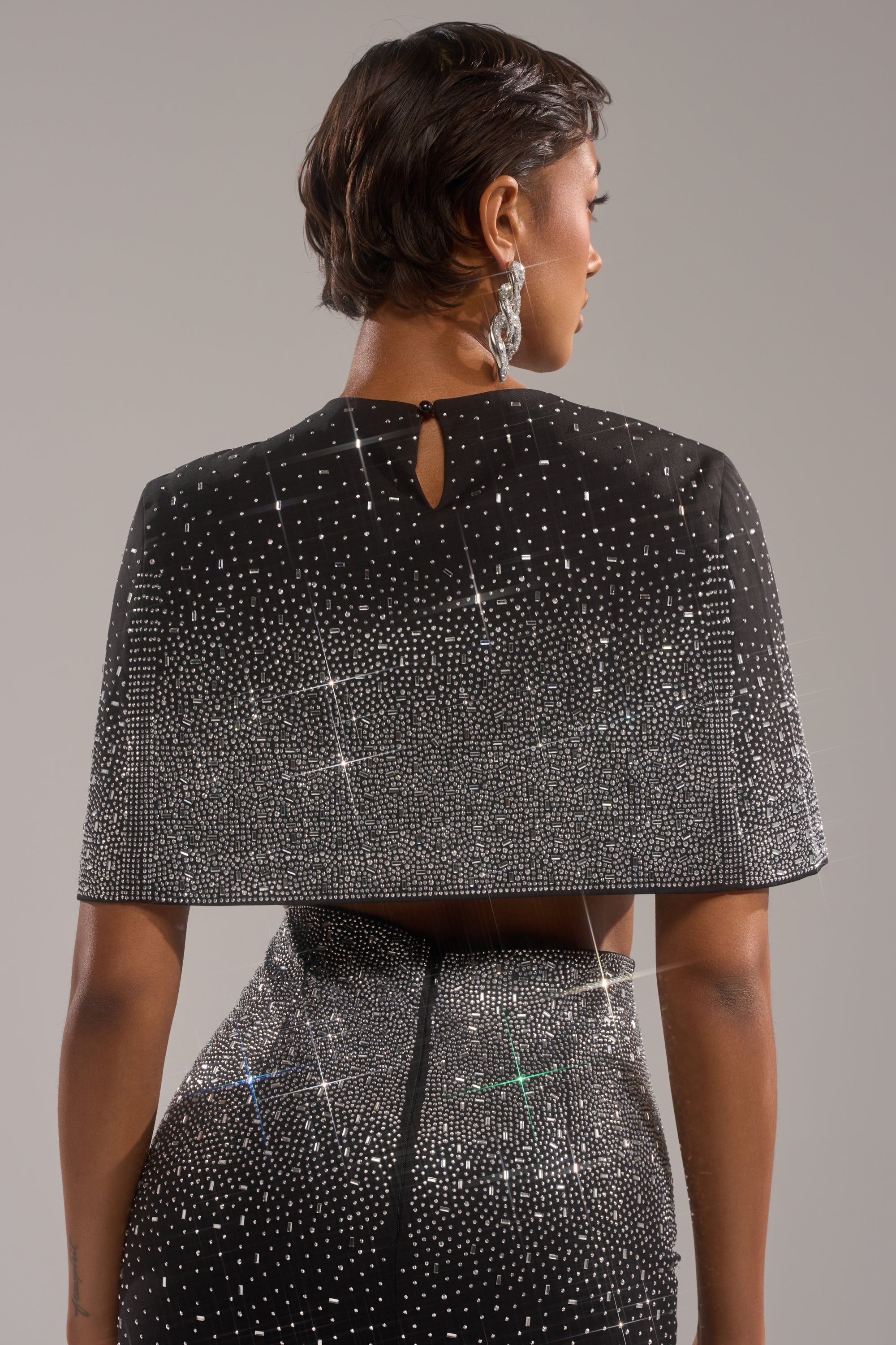 A person with short dark hair, seen from behind, wears the LOVE THIS LIFE BLOUSE—a black capelet-style evening top adorned with sparkling rhinestone embellishments—standing against a neutral background.