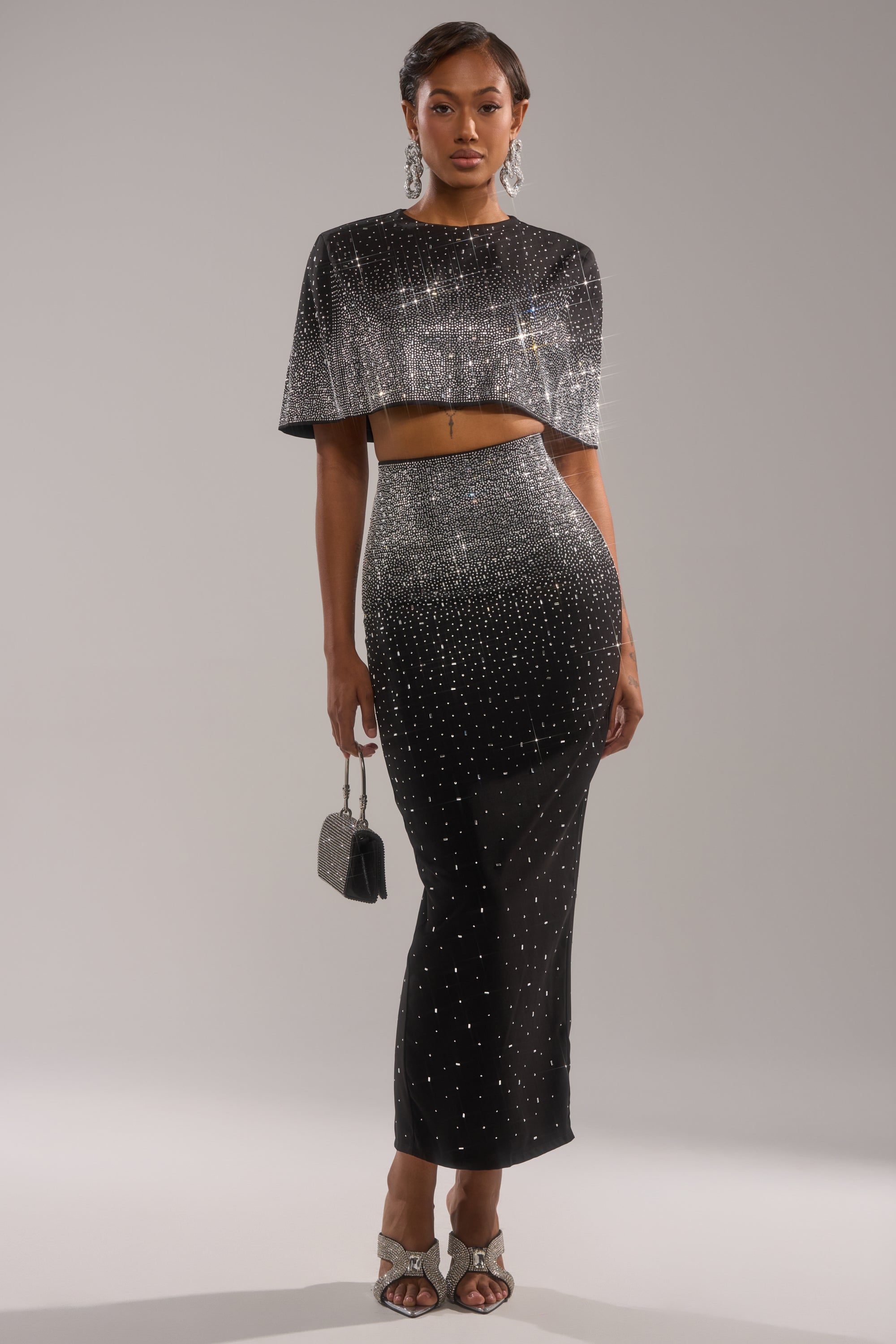 A woman stands against a gray background wearing the LOVE THIS LIFE BLOUSE, a black cropped top with rhinestone embellishments, paired with a matching high-waisted skirt, silver heels, large earrings, and holding a small purse.