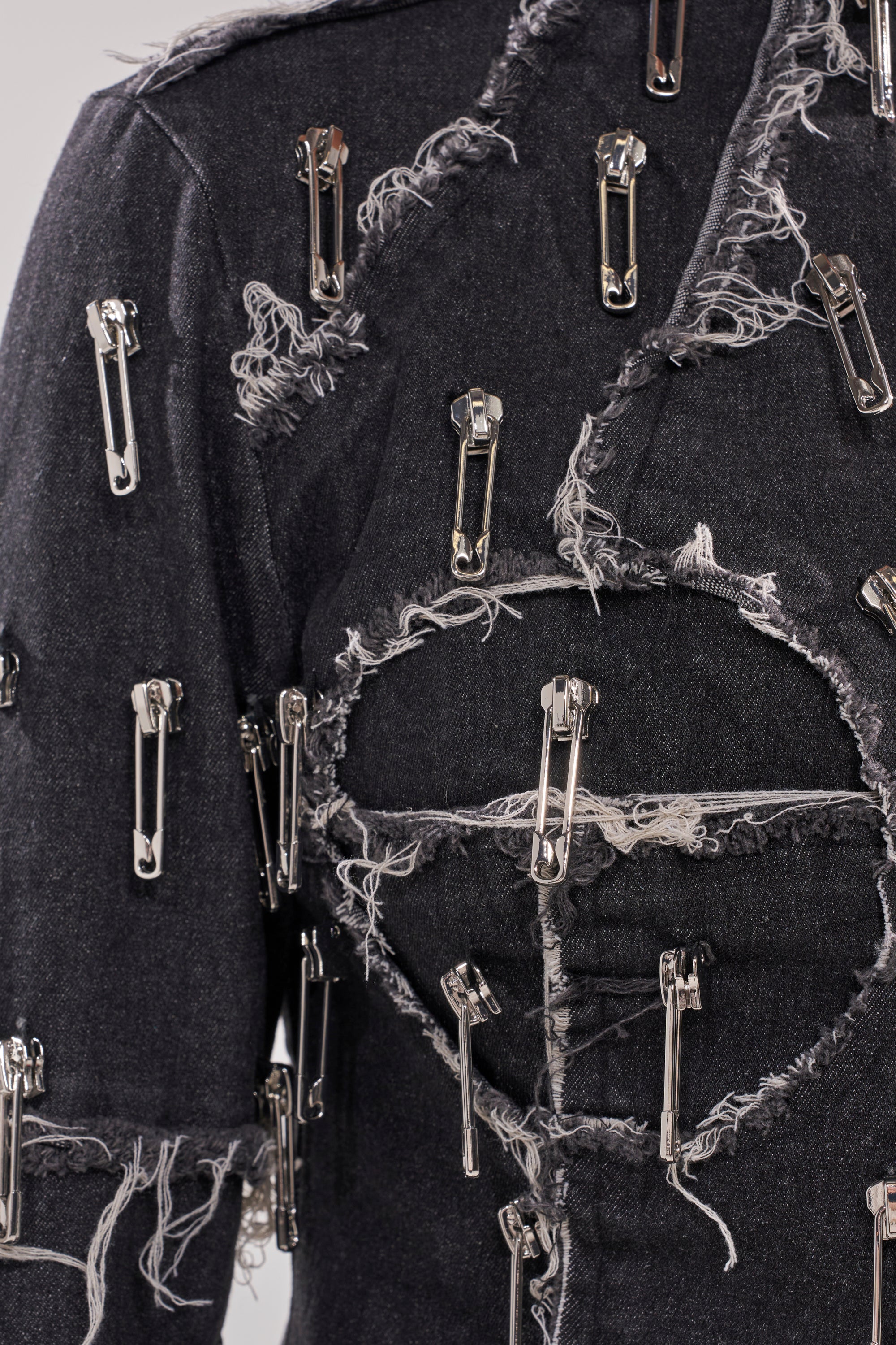 Close-up of the SAINT SERNIN ONE SHOULDER BLOUSE: a dark black denim top, frayed and embellished with silver safety pins arranged in multiple directions for an edgy, avant-garde textured look.