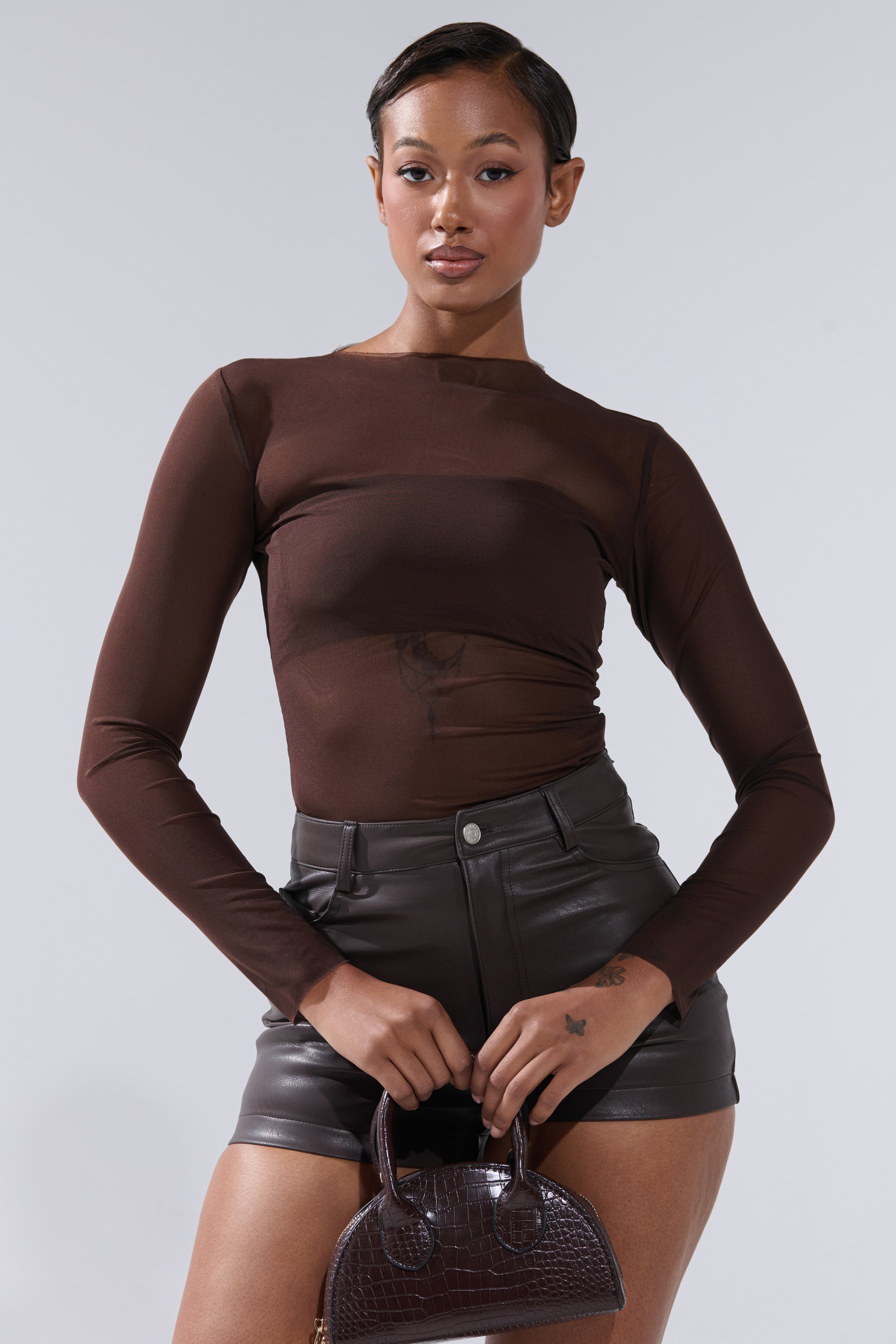 Wearing the Paxton Raw Hem Long Sleeve Mesh Tee in Chocolate Brown with high-waisted black leather shorts, a woman stands holding a small textured brown handbag and looks confidently at the camera against a plain background.