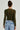 A person with an updo hairstyle is seen from behind, wearing the Paxton Raw Hem Long Sleeve Mesh Tee in Olive and light blue high-waisted jeans with back pockets. The background is plain and light-colored.