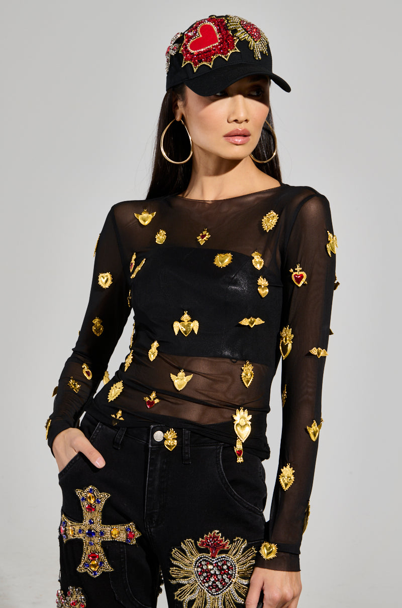 A person wearing the GILDED MESH TOP, embellished with faux gold heart and wing designs, paired with stretchy black jeans featuring ornate embroidery, and a black cap adorned with embroidered heart motifs. They have long hair and are posing with one hand in their pocket.