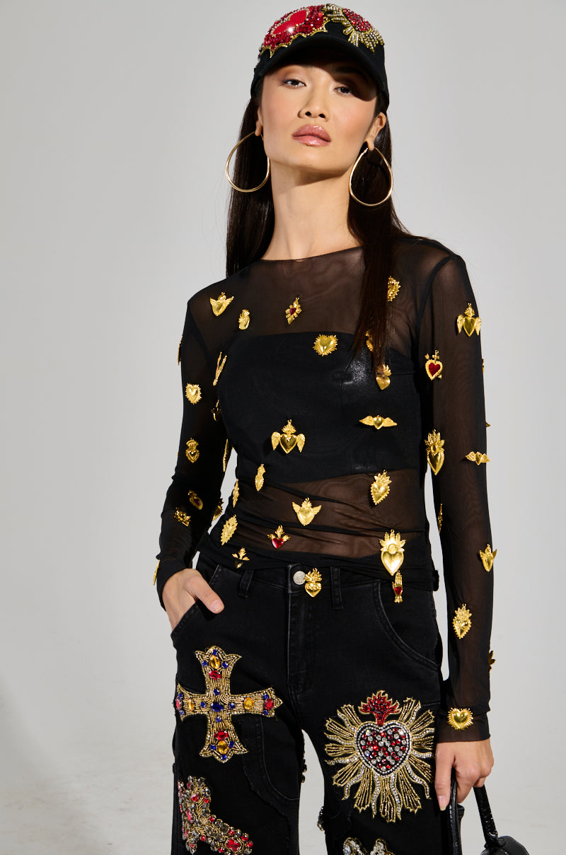 Wearing the GILDED MESH TOP featuring faux gold and red heart-shaped embellishments, a model strikes a confident pose. She completes her look with large hoop earrings, black jeans adorned with decorative patches, and a black cap displaying intricate red and gold designs.