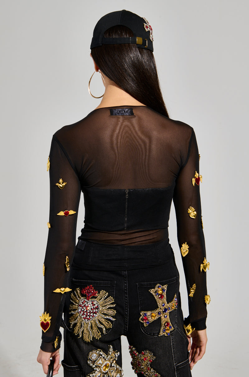 A person is facing away, wearing the GILDED MESH TOP—a black mesh top adorned with faux gold and red embellishments—and black jeans with intricate embroidery. They have long hair, a black cap, and large hoop earrings.