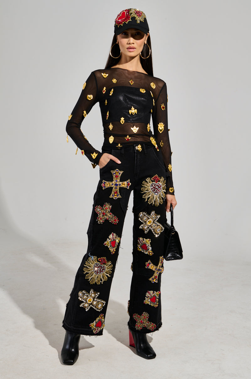 A person dressed in the GILDED MESH TOP, a stretchy and sheer black garment adorned with faux gold embellishments, is paired with black pants that feature ornate cross designs. They complement the look with a matching cap patterned in red, oversized hoop earrings, and carry a stylish black handbag while standing against a plain white background.