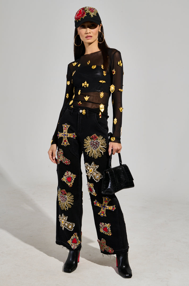A woman poses in a GILDED MESH TOP, which features stretchy black mesh with faux gold embellishments, paired with matching ornate pants. She completes her look with a black cap adorned with a red pattern, large hoop earrings, a chic black handbag, and sleek black boots.