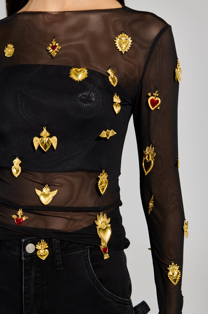 A person wearing a GILDED MESH TOP, featuring stretchy black fabric embellished with faux gold and red heart-shaped pins and symbols, stands with hands out of frame. The top includes long sleeves and is complemented by sleek black pants.