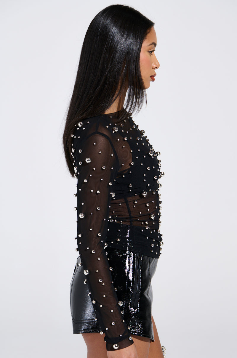 A woman with long black hair is wearing the KYLIE METAL ORB LONG SLEEVE TOP and shiny black shorts, standing in profile against a plain background.