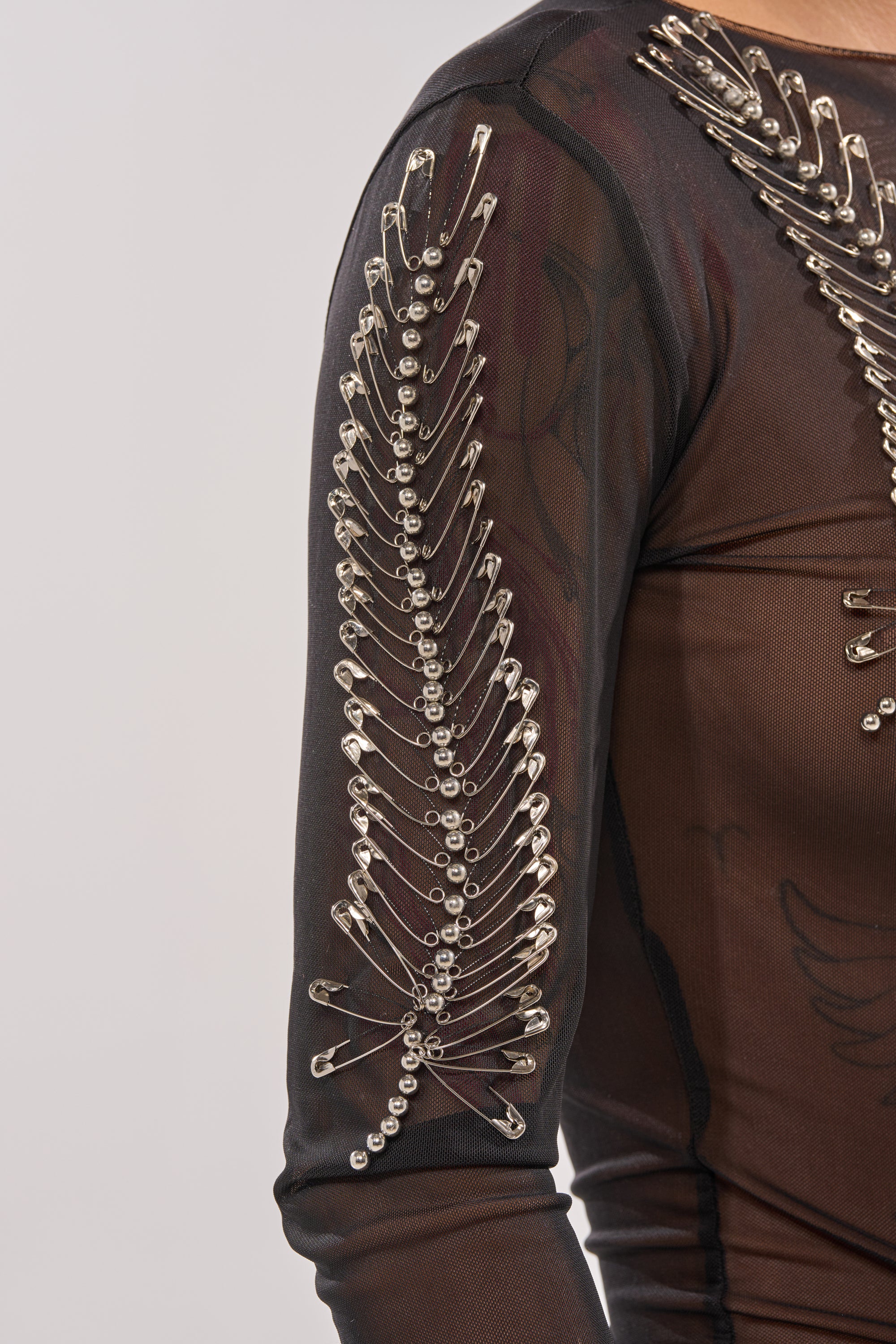 The PINNED DOWN MESH TOP features black sheer mesh with a leaf pattern, adorned with silver safety pin accents and round studs on the shoulder and sleeve—a striking choice for bold nightlife style. 9