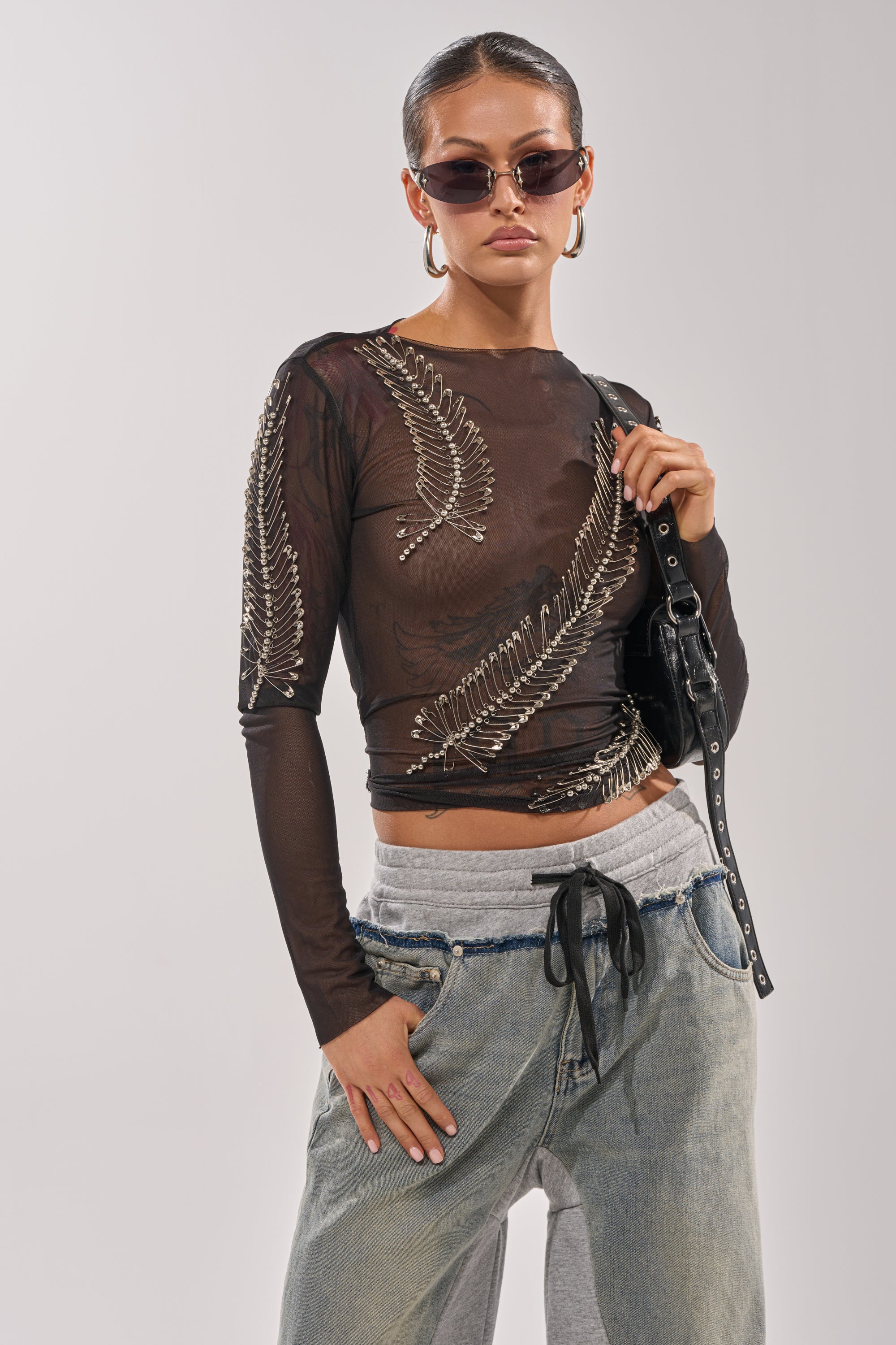 A woman models the PINNED DOWN MESH TOP—black sheer mesh with silver and safety pin details—over a black bra, styled with sunglasses, hoops, and loose jeans layered on gray sweatpants against a plain backdrop; ideal for nightlife fashion. 1