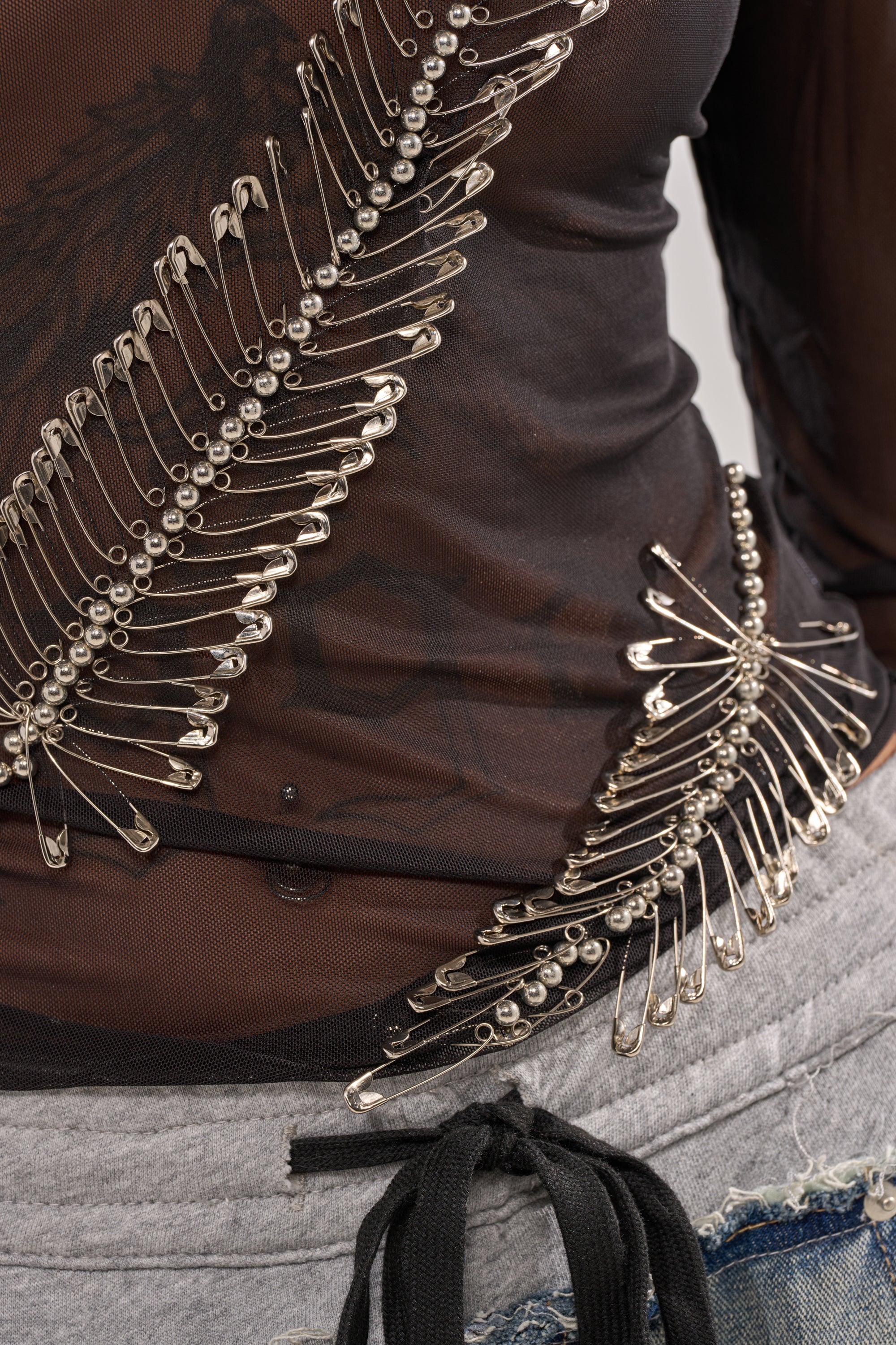 A close-up of a person wearing the PINNED DOWN MESH TOP, a black sheer mesh top with safety pin details, styled with gray sweatpants that feature a black drawstring and frayed blue denim accents for an edgy nightlife-inspired look. 8