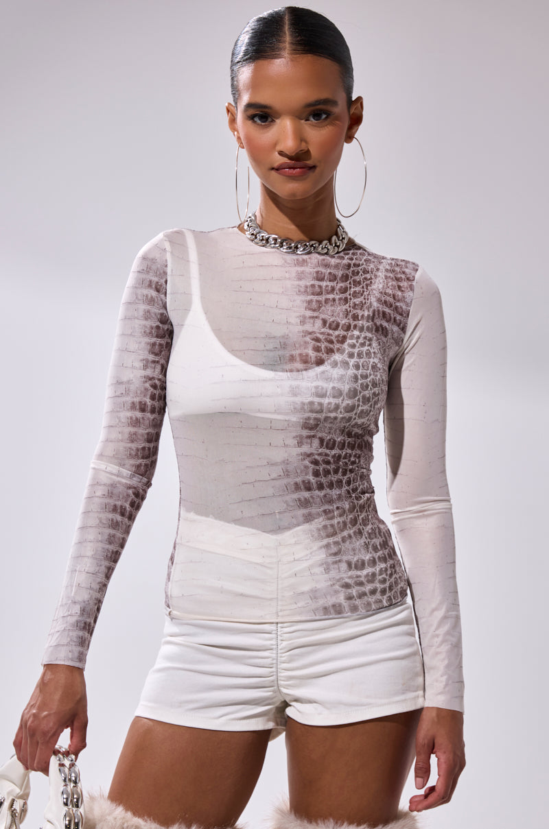 A woman with slicked-back hair wears large hoop earrings, a chunky chain necklace, high-waisted white shorts, and the ELBA PYTHON LONG SLEEVE MESH TOP, standing against a plain background.