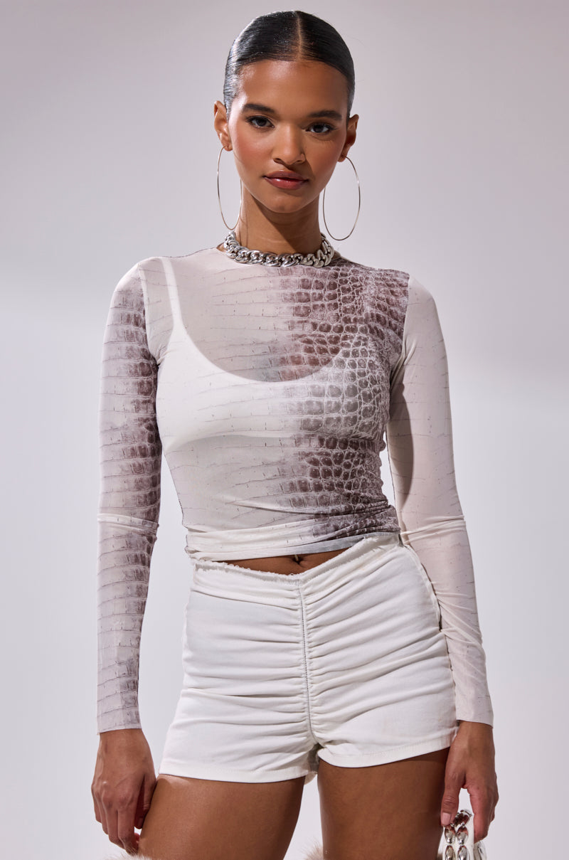 A woman poses confidently against a plain background in the ELBA PYTHON LONG SLEEVE MESH TOP, ruched white shorts, large hoop earrings, and a chunky necklace.