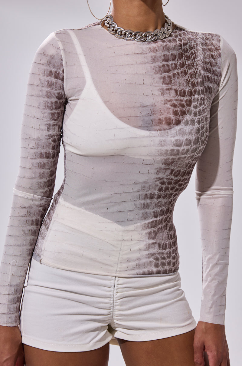 A person wearing the ELBA PYTHON LONG SLEEVE MESH TOP, featuring a fitted mesh design with a python pattern, paired with high-waisted ruched white shorts and a chunky silver chain necklace.