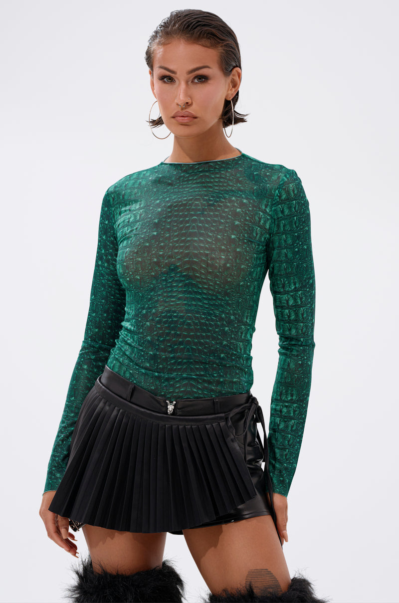 A woman wearing the JESSICA PYTHON LONG SLEEVE MESH TOP—a sheer, green, textured mesh top—paired with a black pleated leather mini skirt and black furry boots stands against a plain white background. 1
