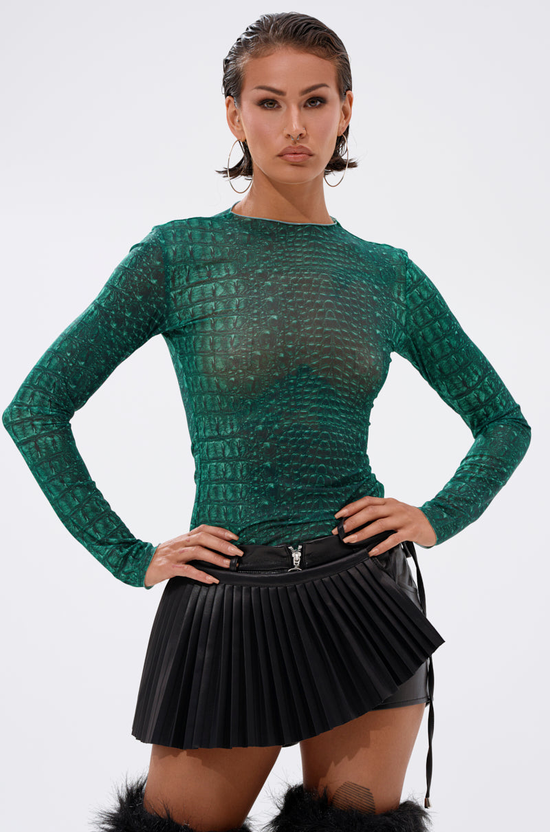 A woman wears the JESSICA PYTHON LONG SLEEVE MESH TOP in emerald green snakeskin with a black pleated mini skirt. She stands hands on hips, gazing confidently at the camera against a plain light background. 0