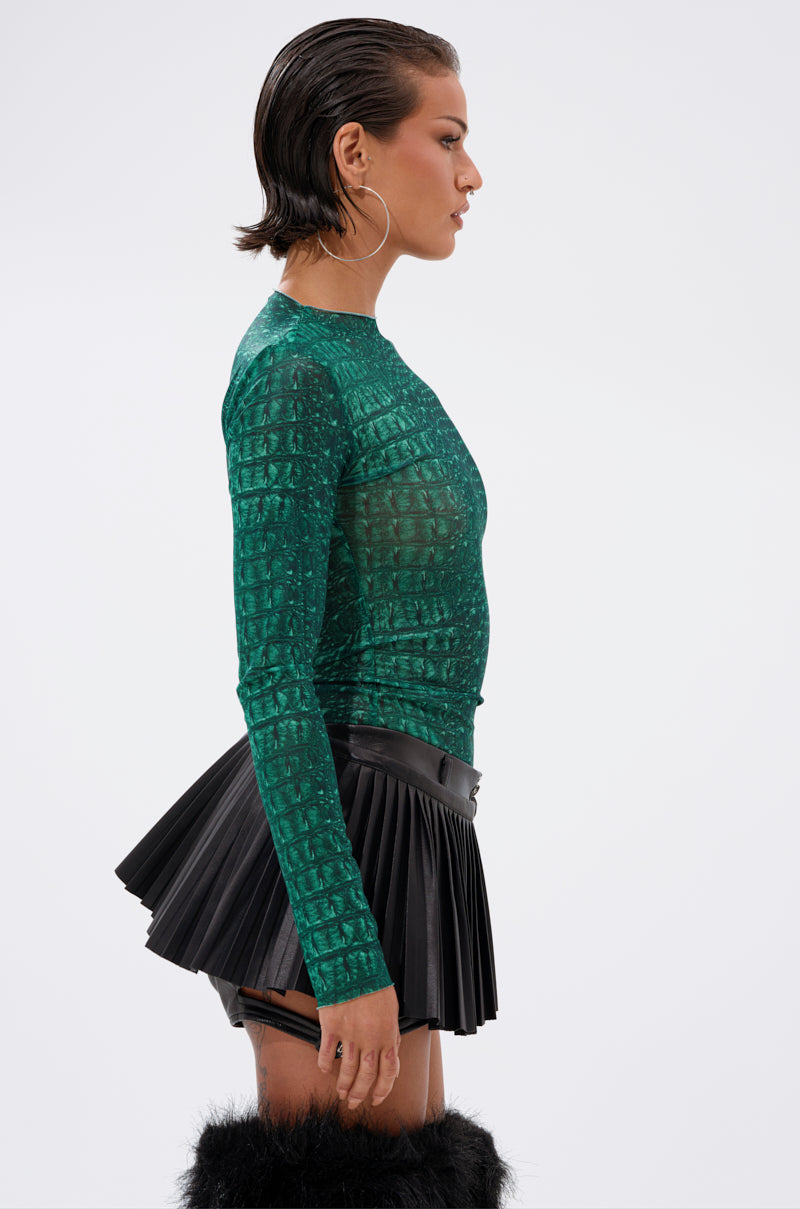 A person with short, slicked-back hair wears the JESSICA PYTHON LONG SLEEVE MESH TOP in emerald green, a black pleated mini skirt, large hoop earrings, and black furry boots, standing in profile against a plain white background. 2