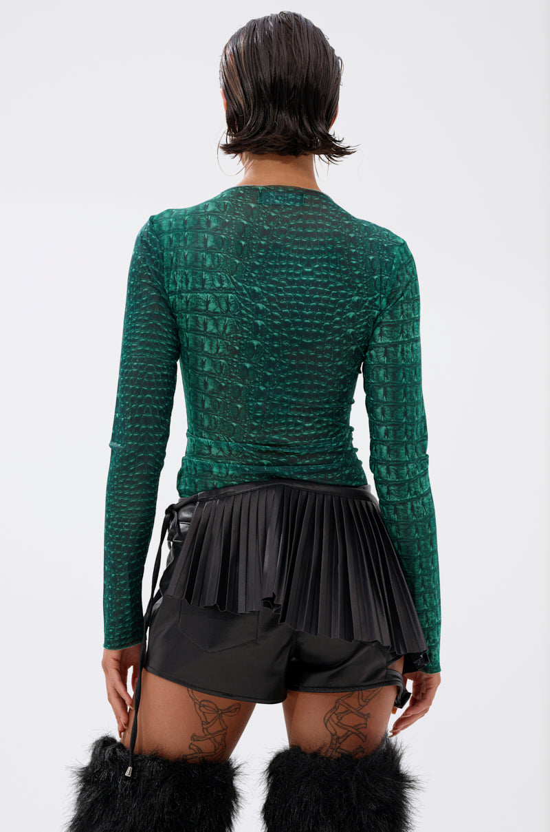 A person with short, wet-look hair is seen from the back wearing the JESSICA PYTHON LONG SLEEVE MESH TOP in emerald green snakeskin, a black pleated mini skirt, black shorts, furry black knee-high boots, and showing thigh tattoos. 3