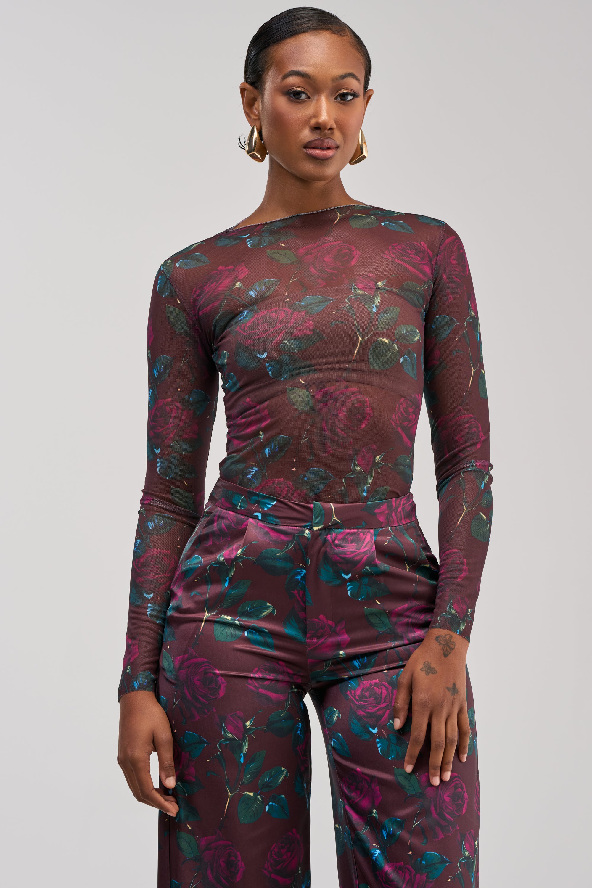 A woman styled with streetwear flair stands against a plain light background, wearing the DOZEN ROSES MESH LONG SLEEVE TOP IN BURGUNDY paired with high-waisted pants in coordinating burgundy, teal, and green tones.