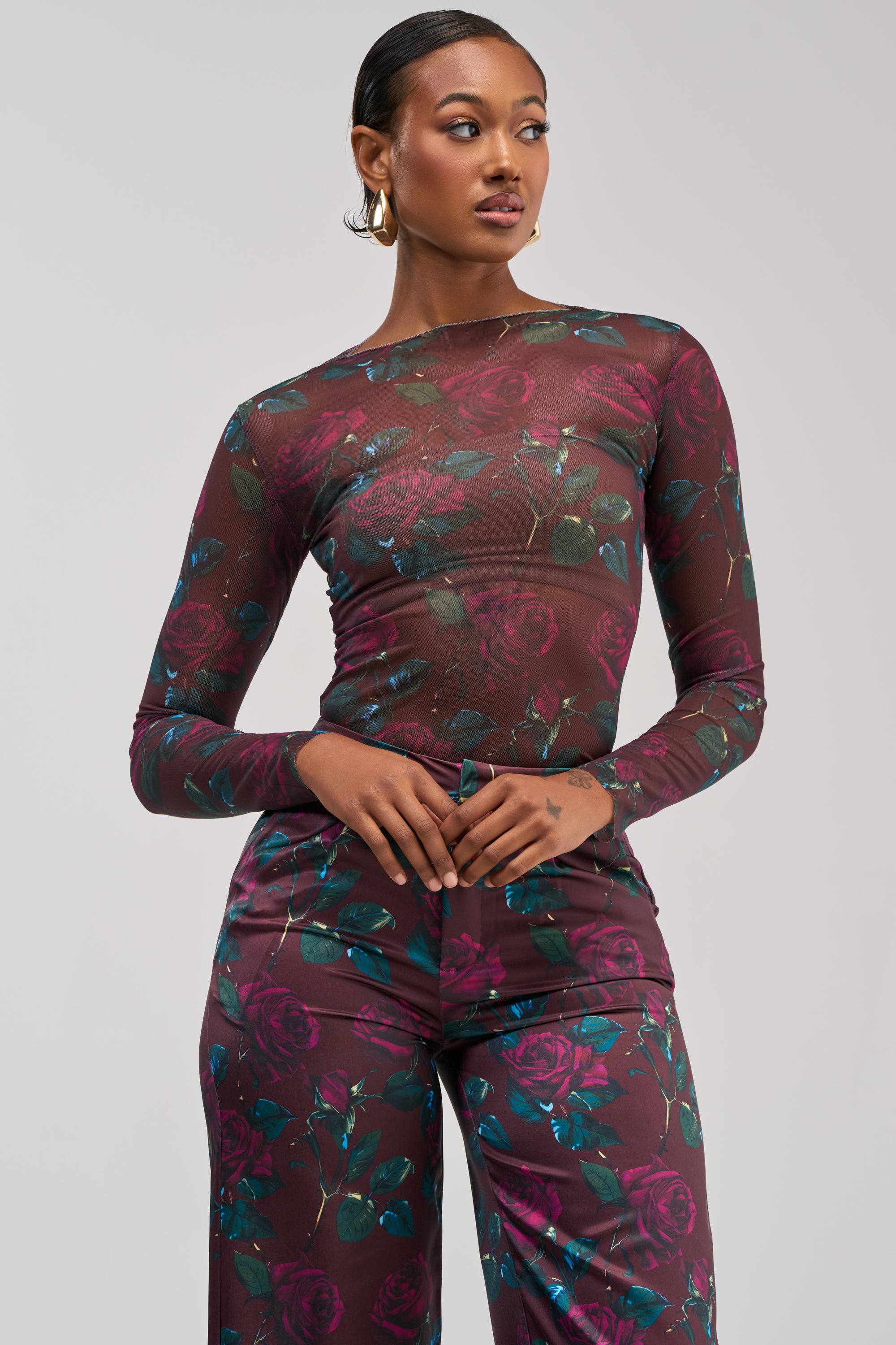 A woman wearing the DOZEN ROSES MESH LONG SLEEVE TOP IN BURGUNDY with matching pants featuring a dark floral pattern stands against a light gray background, looking to her left with her hands clasped at her waist.