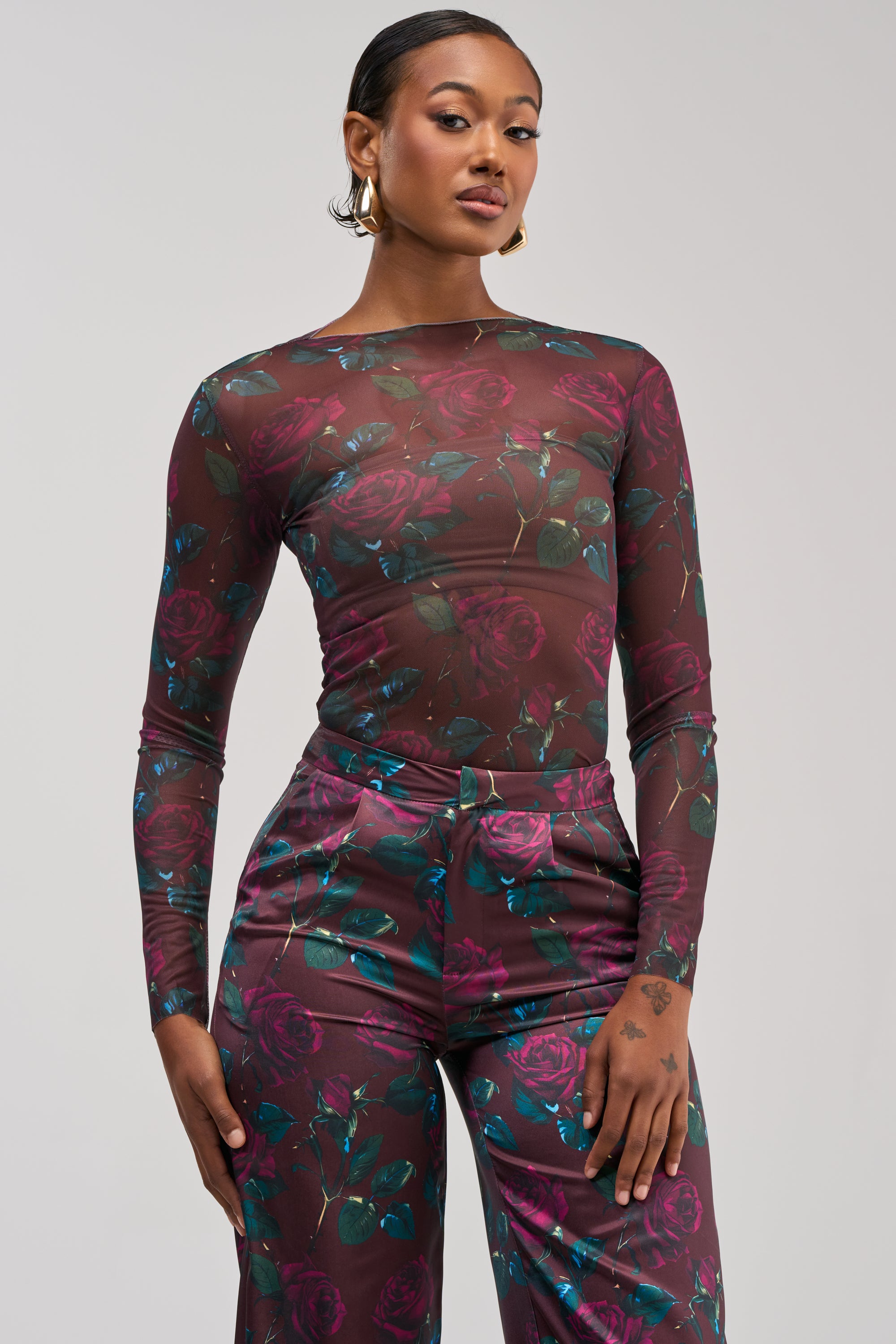 A woman with short hair, gold hoop earrings, and neutral makeup strikes a confident pose against a light gray background, wearing the DOZEN ROSES MESH LONG SLEEVE TOP IN BURGUNDY and matching rose print pants for an elevated streetwear look.