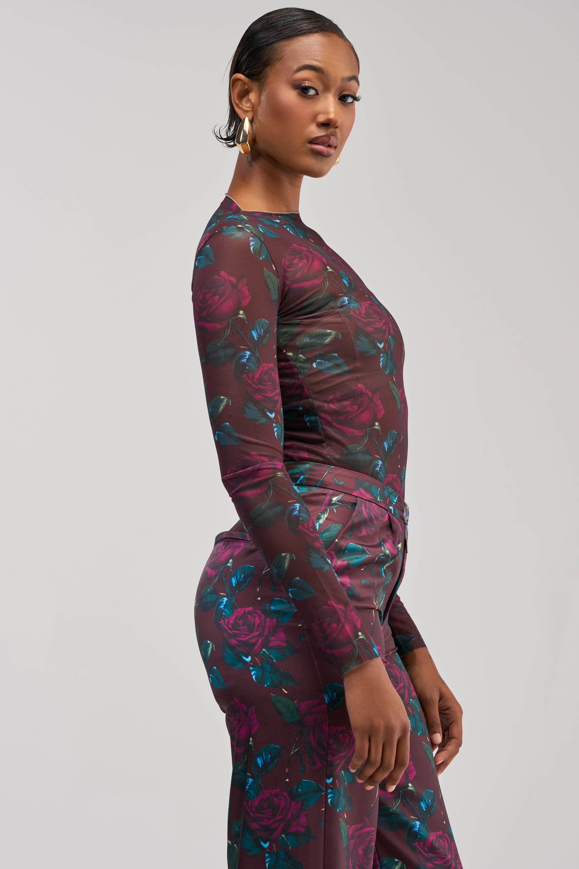 A woman with short, slicked-back hair and gold hoop earrings poses in profile against a light gray background, channeling effortless streetwear style in the DOZEN ROSES MESH LONG SLEEVE TOP IN BURGUNDY paired with matching dark floral print pants.