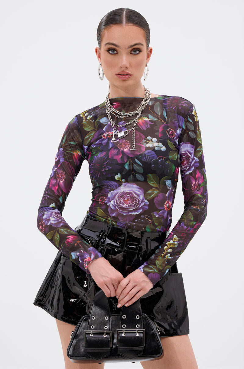 A woman with slicked-back hair wears the ANOTHER ROSE LONG SLEEVE MESH TOP, layered necklaces, hoop earrings, a shiny black mini skirt, and holds a small black handbag in front of a plain white background. 0