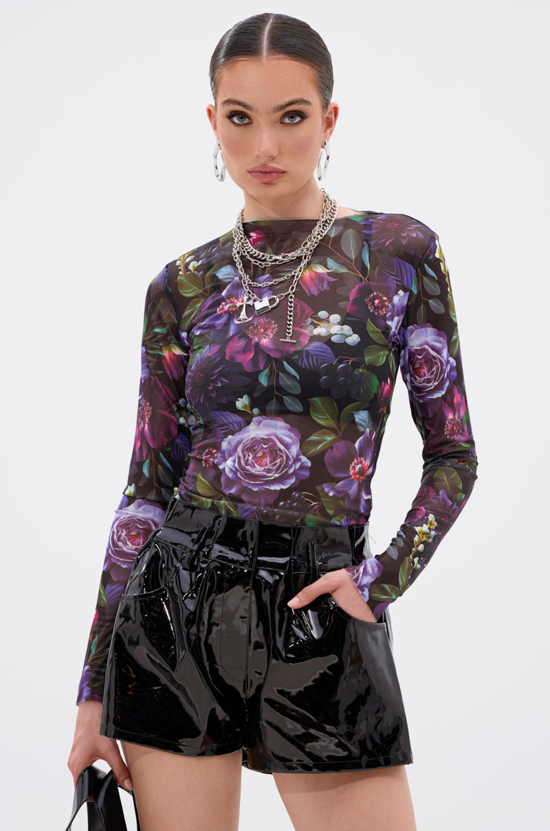 A woman with sleek hair and bold makeup wears the ANOTHER ROSE LONG SLEEVE MESH TOP, paired with shiny black high-waisted shorts, layered silver necklaces, and hoop earrings. She poses confidently with one hand in her pocket. 1