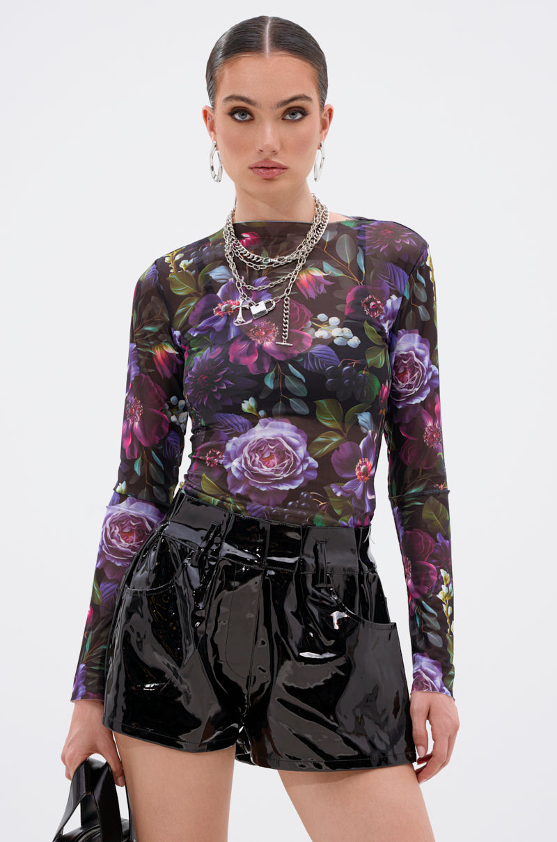 She wears the ANOTHER ROSE LONG SLEEVE MESH TOP with shiny black vinyl shorts, hoop earrings, and layered chain necklaces. With slicked-back hair and black shoes in hand, she poses confidently against a plain white background. 2