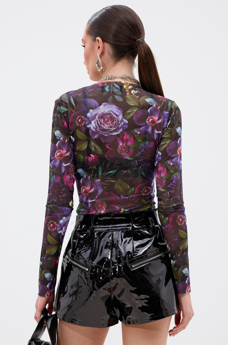 A woman with a sleek ponytail wears the ANOTHER ROSE LONG SLEEVE MESH TOP and shiny black vinyl shorts. She faces away, holding a black handbag against a white background. 4