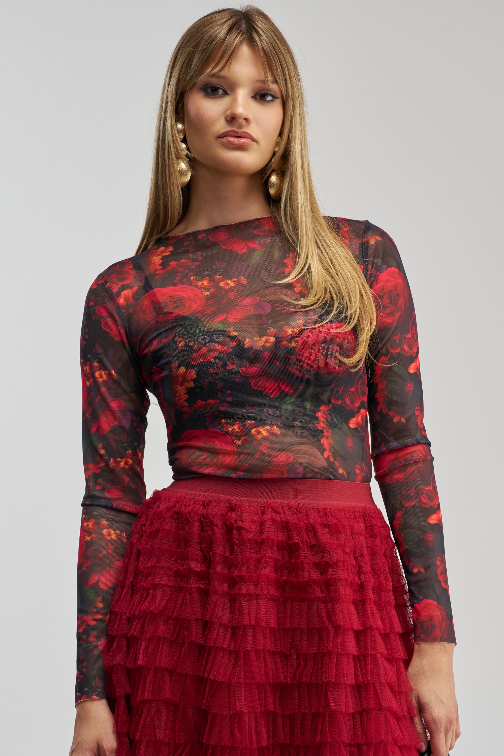 A woman wears the ROSE GARDEN LONG SLEEVE MESH TEE, featuring red and green floral patterns, paired with a red layered ruffle skirt. She has straight light brown hair, large gold earrings, and stands against a plain gray background.