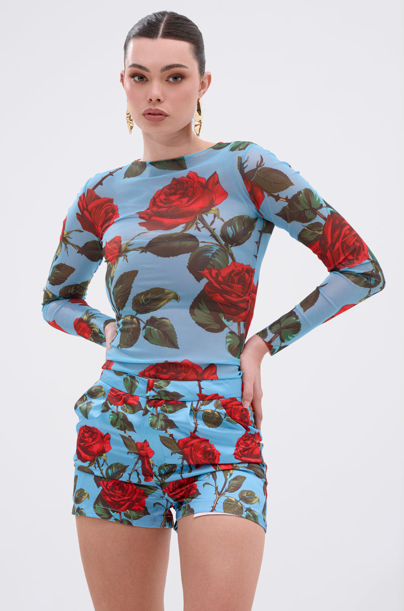 A woman poses confidently in the DOZEN ROSES MESH TOP, a blue long-sleeve mesh shirt with a vibrant floral print, paired with matching shorts and gold earrings, set against a plain white background.