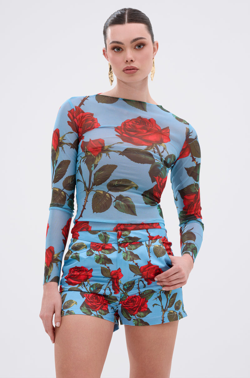 A woman stands against a plain white backdrop wearing the DOZEN ROSES MESH TOP, which features mesh fabrication and a bold red rose floral print on a blue background, paired with high-waisted shorts and one hand in her pocket.