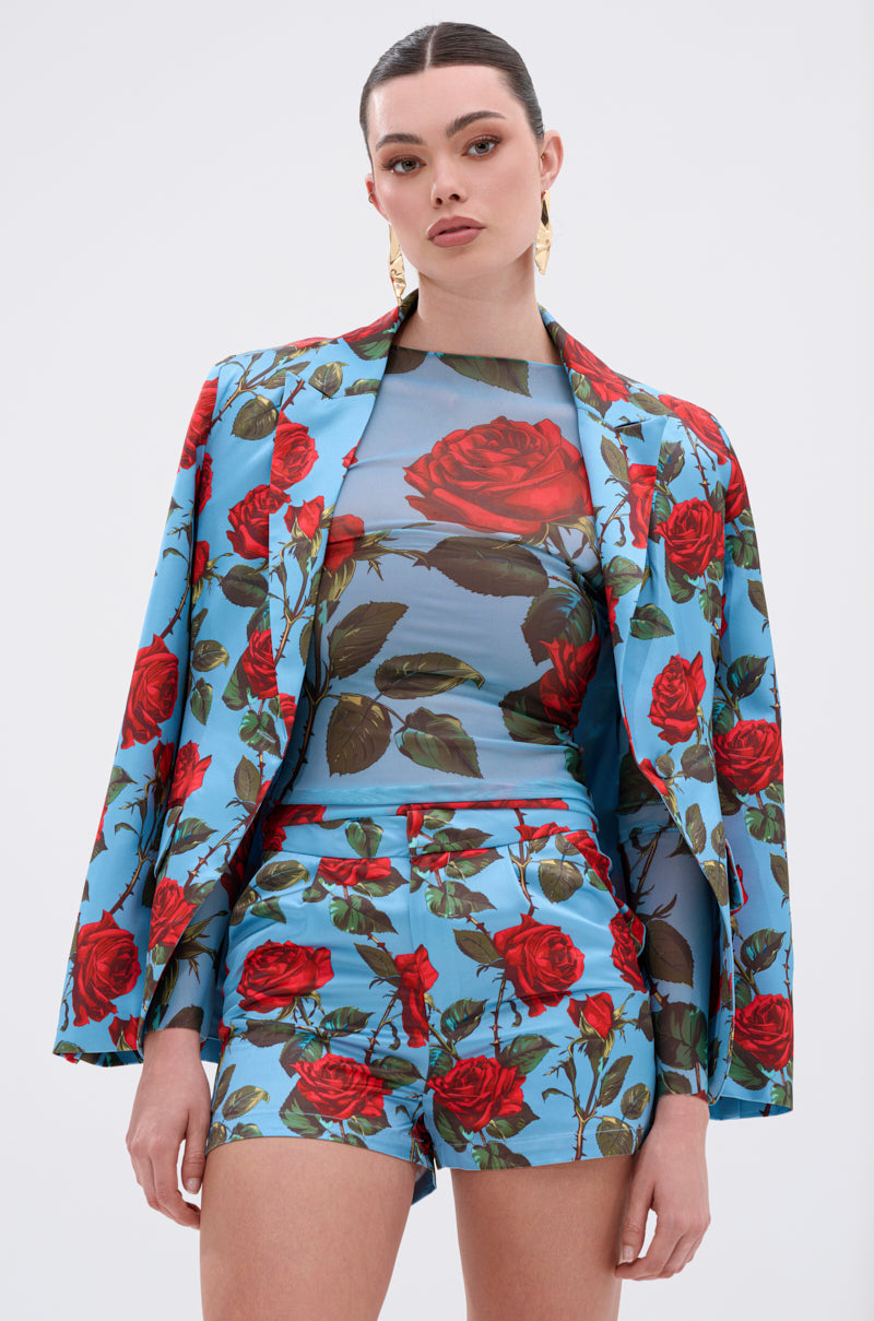A woman wears the DOZEN ROSES MESH TOP as part of a bold blue floral ensemble, including a blazer over her shoulders and high-waisted shorts. She styles her hair back and accessorizes with gold earrings.