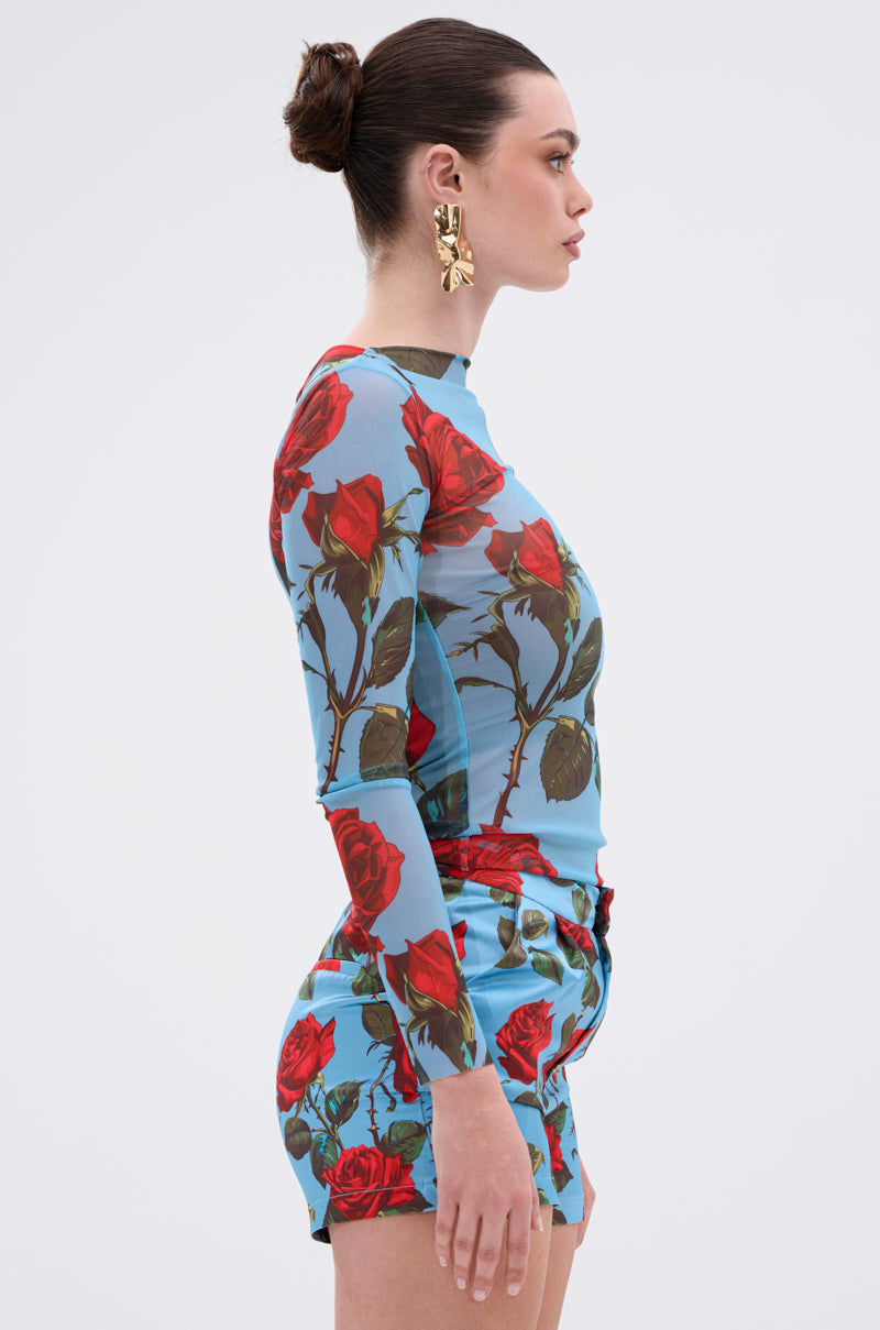 A woman with a bun and large gold earrings wears the DOZEN ROSES MESH TOP featuring mesh fabric and a bold floral graphic, paired with matching shorts, as she stands in profile against a plain light background.