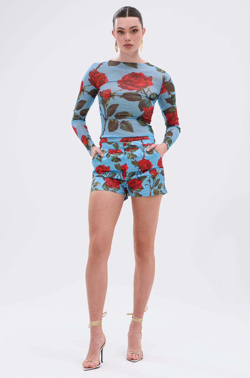 A woman faces forward in the DOZEN ROSES MESH TOP and matching shorts, both featuring a bold blue floral graphic. Her hands are in her pockets as she wears strappy high-heeled sandals, set against a plain white background.