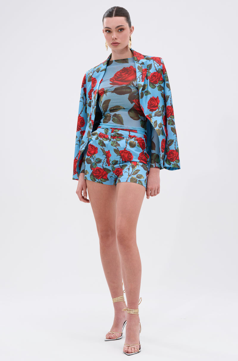 A woman poses against a white background in a blue jacket and shorts with a red floral print, the DOZEN ROSES MESH TOP, gold hoop earrings, and strappy high-heeled sandals.