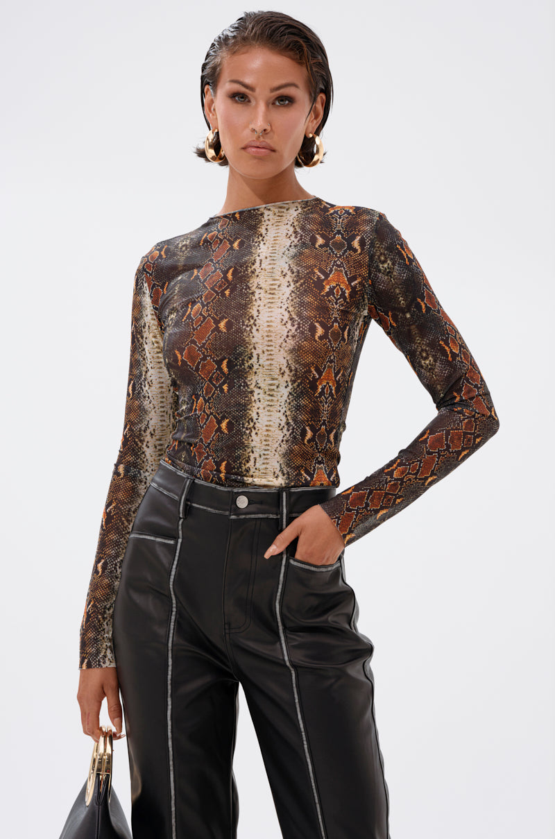 A woman stands confidently in the SNAKE VIBES MESH TOP featuring a brown and tan snakeskin print, styled with high-waisted black leather pants, a black handbag, slicked-back hair, gold hoop earrings, and a neutral expression.