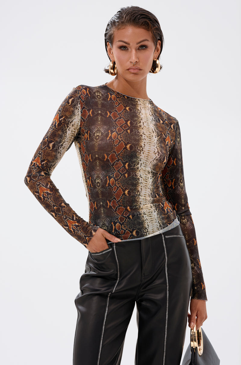A woman with wet-look hair wears the SNAKE VIBES MESH TOP with sheer long sleeves, black leather pants featuring white stitching, gold hoop earrings, and holds a black bag with a gold handle against a plain background.