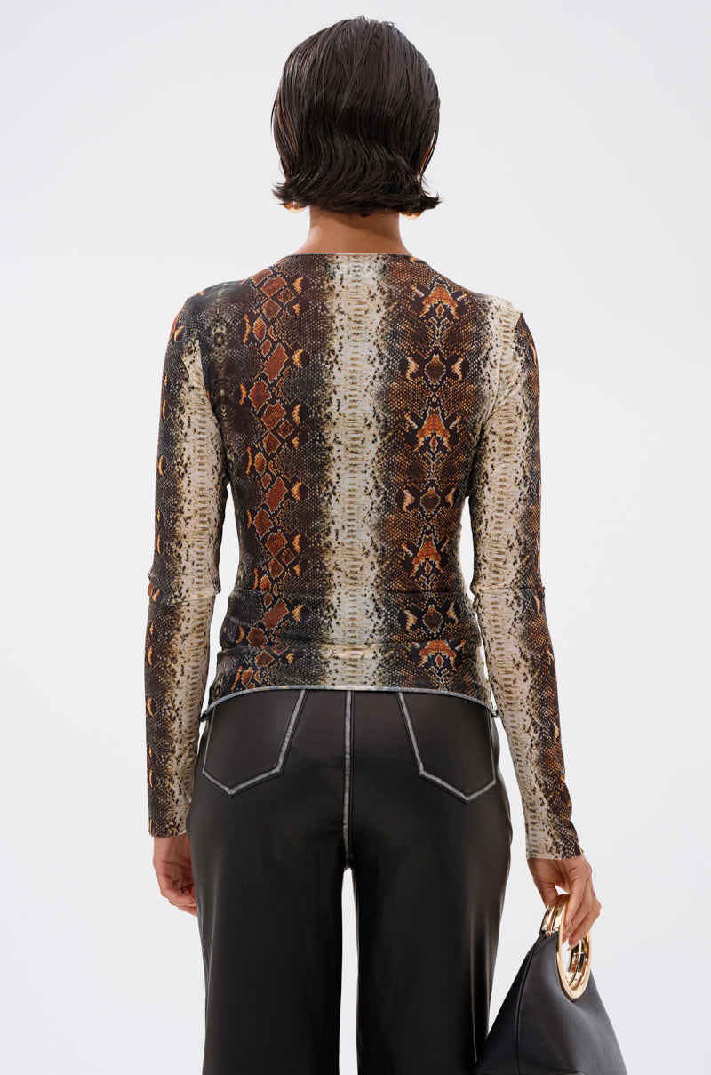 A person with short dark hair, seen from behind, wears the SNAKE VIBES MESH TOP—long-sleeved with a brown and beige snakeskin print—paired with black pants with white stitching and a black handbag featuring a gold handle.