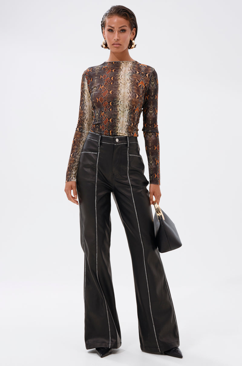 A woman stands against a plain background wearing the SNAKE VIBES MESH TOP, high-waisted black leather pants with white stitching, pointed shoes, and holding a black handbag.